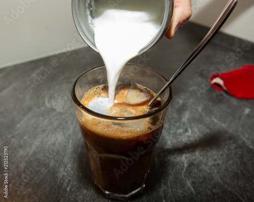 Person is pouring frothed milk into a glass filled with iced coffee and a long spoon.