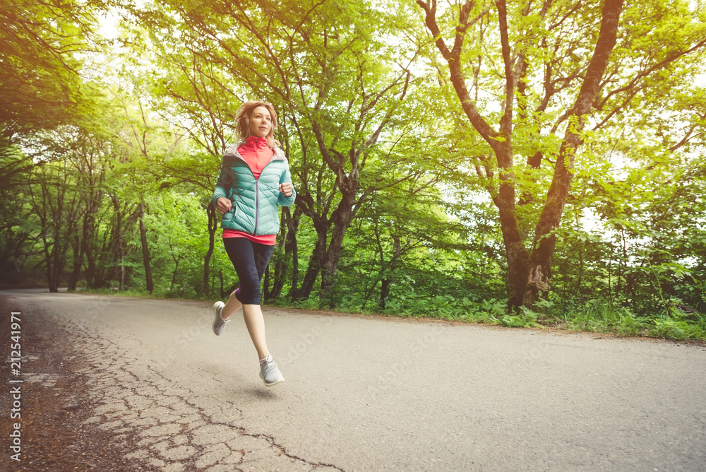 Young fitness blonde woman in headphones running at morning caucasian forest trail in sun light Young fitness blonde woman in headphones running at morning caucasian forest trail in sun light