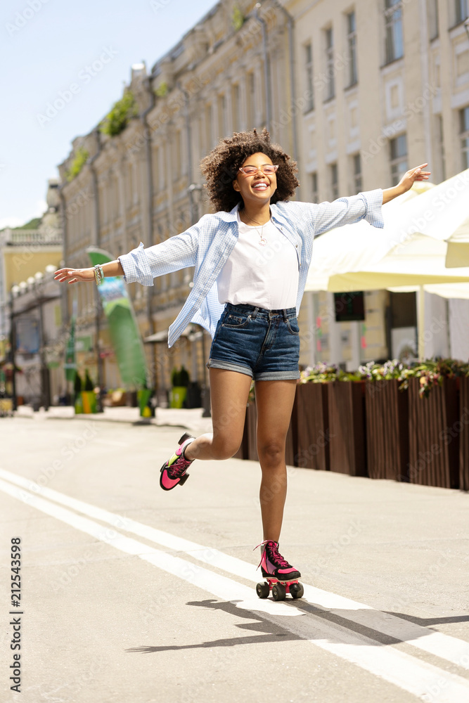 Foto de Enjoying herself. Pretty curly girl enjoying roller-skating down  the street and spreading her hands wide while smiling do Stock