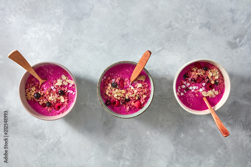 Dietary breakfast of beetroot smoothies with raspberries, currants, granola and sesame seeds in a plate with a wooden spoon on a gray concrete background. Top view