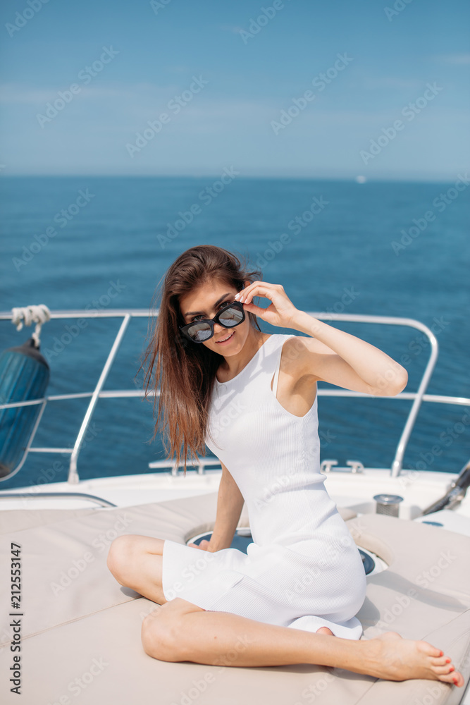 Outdoor shot of adorable young woman in white elegant dress sitting on edge of boat and looking at camera during sea trip. Happy woman enjoying summer travel. Vacation or holiday concept