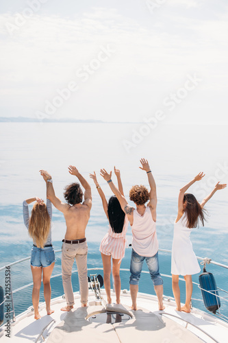 Back view of celebrating young elite rich teeenagers standing on the bow of deck of comfortable yacht with hands raised up, being happy with luxury and carefree life.