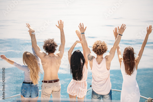 Rear view of cheerful caucasian teenagers on a sailing boat, lifting their hands up, looking at blue sea surfice, full of happiness,celebrating the start of their summer vacations.