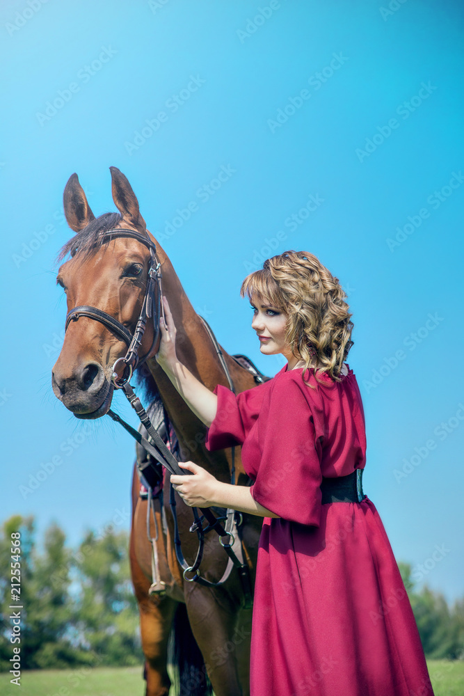 Obraz premium Beautiful girl in a long red dress stands next to the brown horse. A girl stroking a horse.