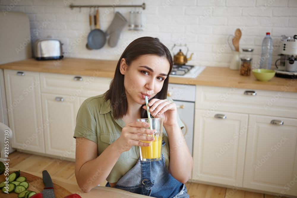 Portrait of cheerful young woman with chubby cheeks and dark hair having fresh orange juice for breakfast. Pretty brunette girl sipping smoothie through straw, siting in modern kitchen interior Portrait of cheerful young woman with chubby cheeks and dark hair having fresh orange juice for breakfast. Pretty brunette girl sipping smoothie through straw, siting in modern kitchen interior