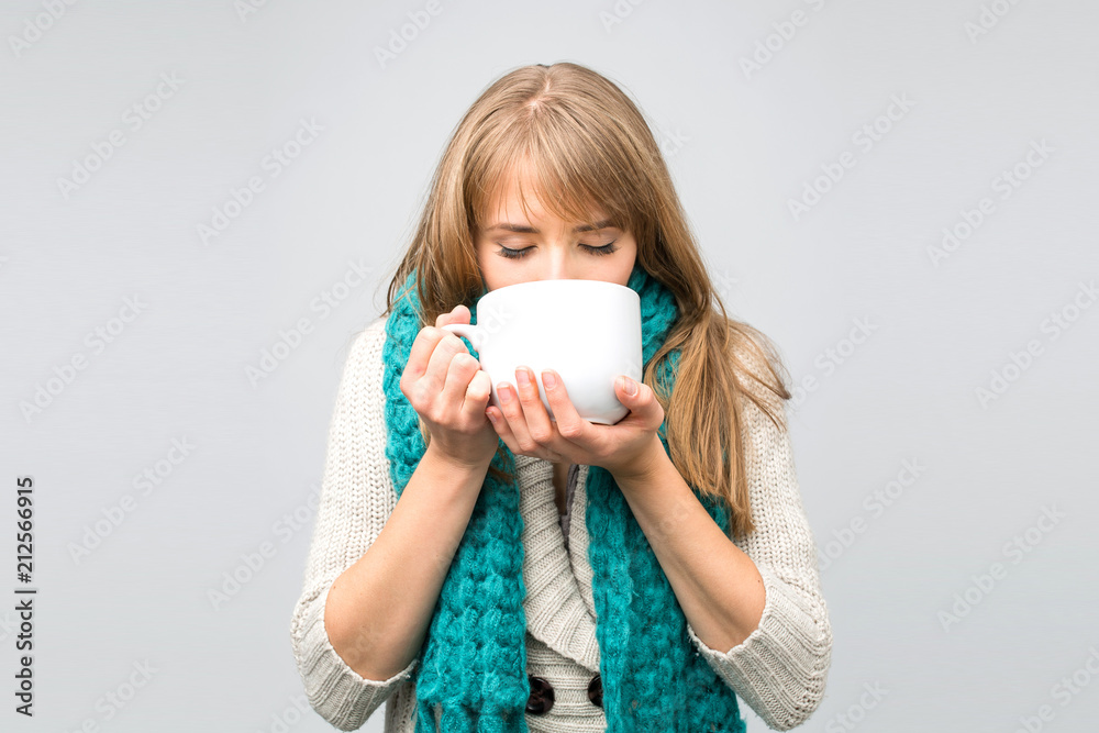 Pensive young woman in knitted cap and blue scarf drinking tea from big cup standing against grey wall . Pensive young woman in knitted cap and blue scarf drinking tea from big cup standing against grey wall .