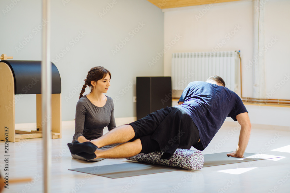 Fototapeta premium Female coach rehabilitation specialist helping young handsome man doing exercise with side surface shin. Male feeling pain while using foam roller in pilates studio. Rehabilitation after trauma.