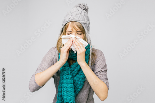 Young woman in knitted cap blowing her nose with pocket tissue, grey wall in background .