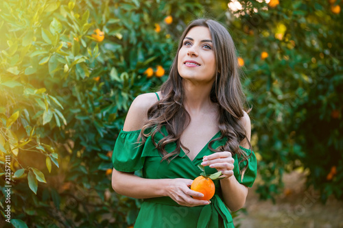 Young woman  outdoors at sunset in a orange orchard  looking at camera and smiling  holding an orange fruit. Healthy lifestyle concept  skin and hair care concept.