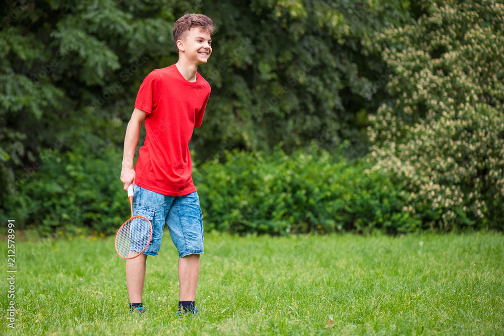 Young teen boy playing badminton in meadow with forest in background. Child with badminton rackets in hand. Kid have fun in summer park at day. Young teen boy playing badminton in meadow with forest in background. Child with badminton rackets in hand. Kid have fun in summer park at day.