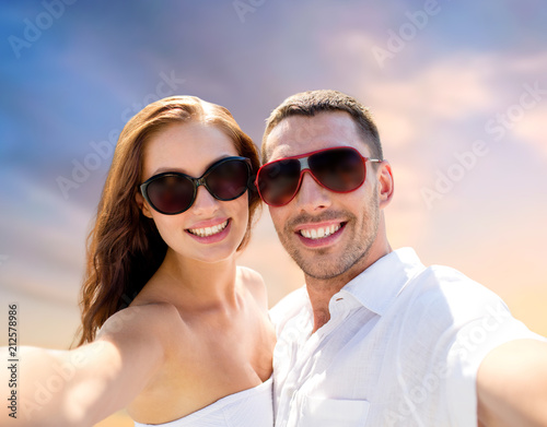love, summer and people concept - smiling couple wearing sunglasses making selfie over evening sky background