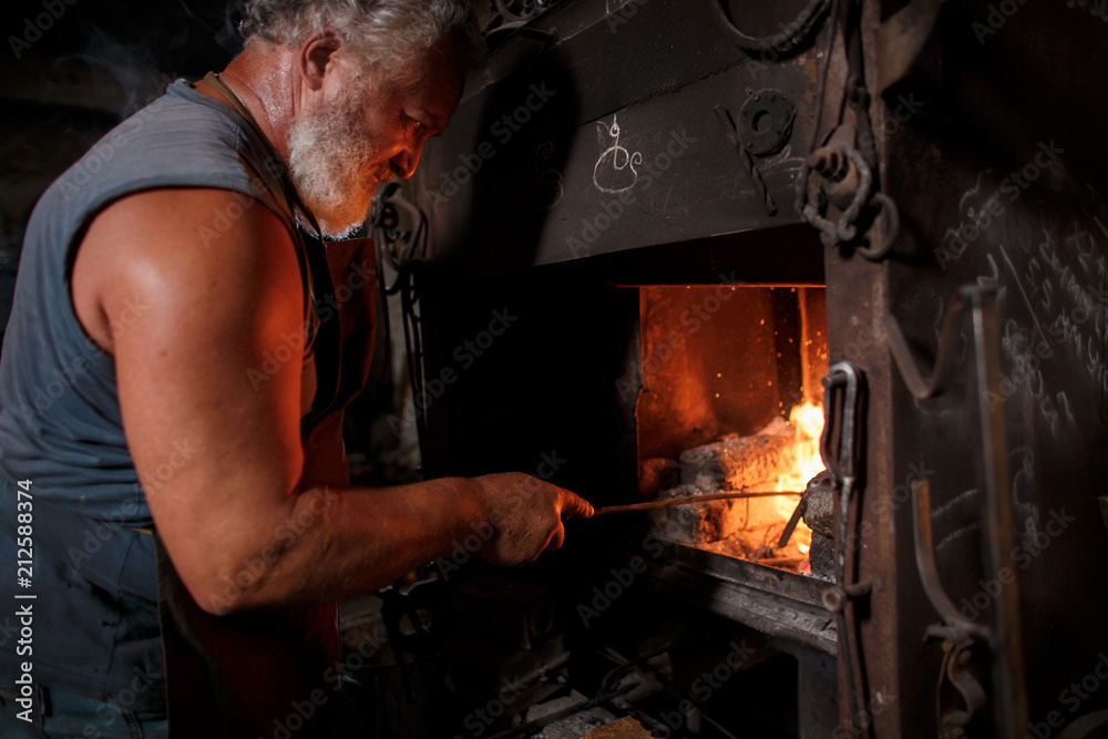The smith artisan in his apron heats the workpiece in the burning furnace in the smith The smith artisan in his apron heats the workpiece in the burning furnace in the smith