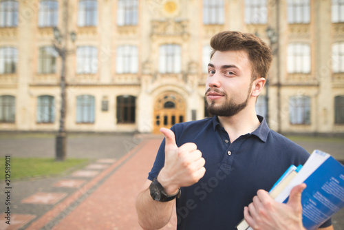 Portrait of a handsome student standing with books in the hands of the university building background and showing a thumbs up. Happy man student on campus university.Student likes the university. photo