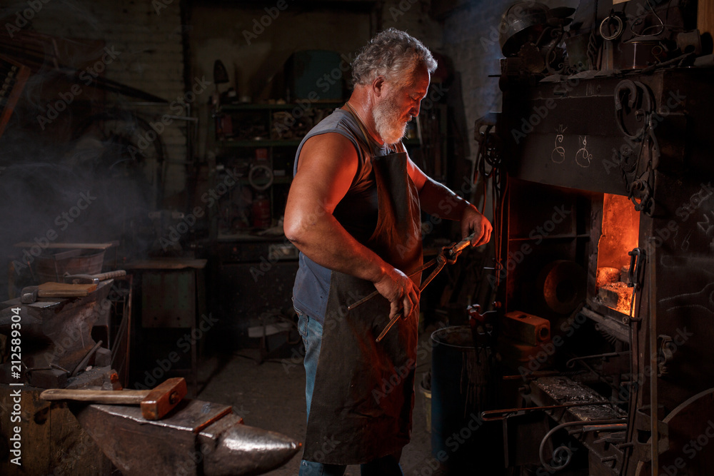 The smith artisan in his apron heats the workpiece in the burning furnace in the smith The smith artisan in his apron heats the workpiece in the burning furnace in the smith