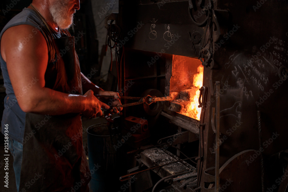 The smith artisan in his apron heats the workpiece in the burning furnace in the smith The smith artisan in his apron heats the workpiece in the burning furnace in the smith
