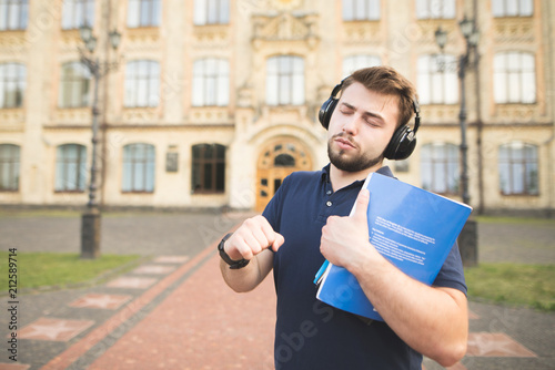 Portrait of a man standing on the background of the university building with books in his hands and listening to music in the headphones. Student with headphones on the campus of the university. photo