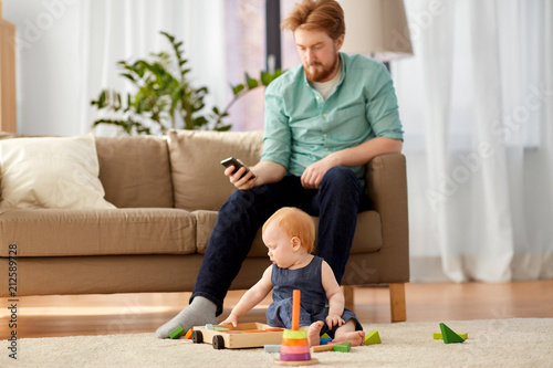 family, fatherhood and people concept - happy red haired father with smartphone and little baby daughter playing with toy blocks kit at home