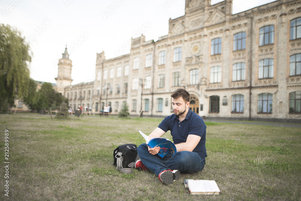 Portrait of a student sitting on the grass and reading a book on the background of a beautiful old university. Man studying outdoors on the campus of the University