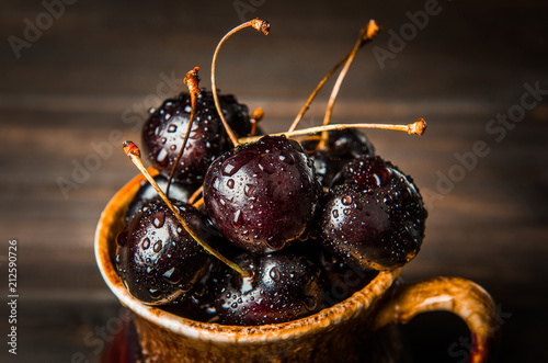 Ripe sweet cherry berries with drops in a brown earthenware plate on a wooden surface background.
