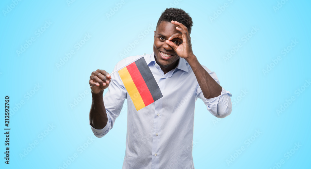Young african american man holding german flag with happy face smiling doing ok sign with hand on eye looking through fingers Young african american man holding german flag with happy face smiling doing ok sign with hand on eye looking through fingers
