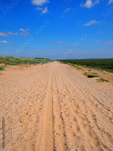 a straight long dry sand road with tire tracks and footprints extending to the horizon surrounded by grass covered dunes with a bright blue summer sky