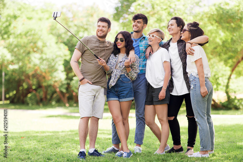 Fototapeta Naklejka Na Ścianę i Meble -  friendship, leisure and technology concept - group of happy smiling friends taking picture by selfie stick at summer park