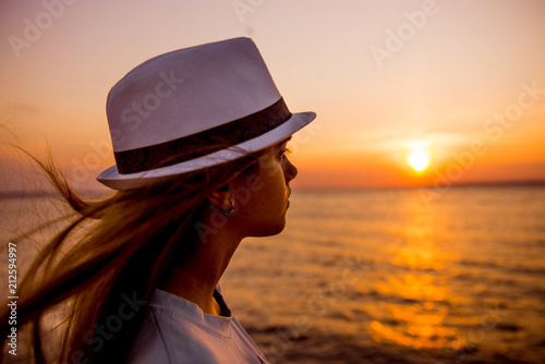 Young girl on the beach at sunset. A girl in a white hat on the beach in the evening. Woman tourist looking at sunset at sea 