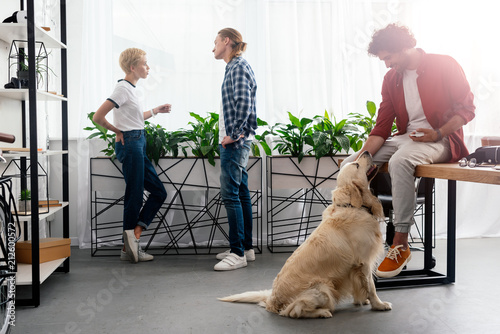 young man stroking dog while colleagues talking near window in office