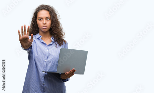 Young hispanic woman holding computer laptop with open hand doing stop sign with serious and confident expression, defense gesture