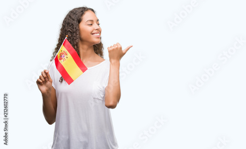 Young hispanic woman holding flag of Spain pointing and showing with thumb up to the side with happy face smiling photo