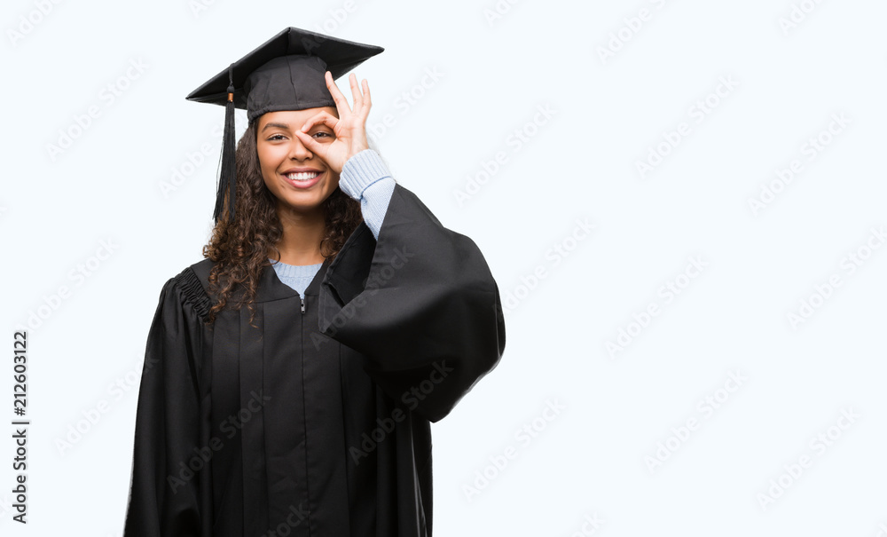 Young hispanic woman wearing graduation uniform with happy face smiling doing ok sign with hand on eye looking through fingers Young hispanic woman wearing graduation uniform with happy face smiling doing ok sign with hand on eye looking through fingers
