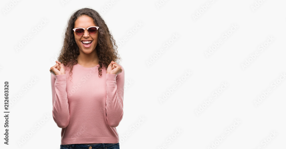 Beautiful young hispanic woman wearing sunglasses excited for success with arms raised celebrating victory smiling. Winner concept.
