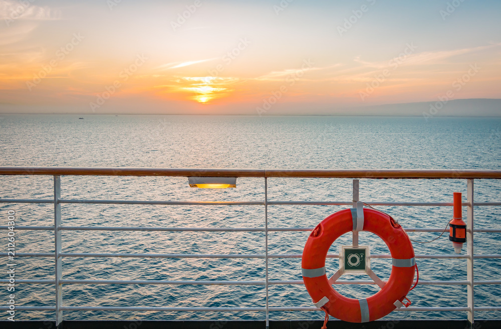 Red lifebuoy on safety railing of cruise ship. Sunset at the ocean in the background.