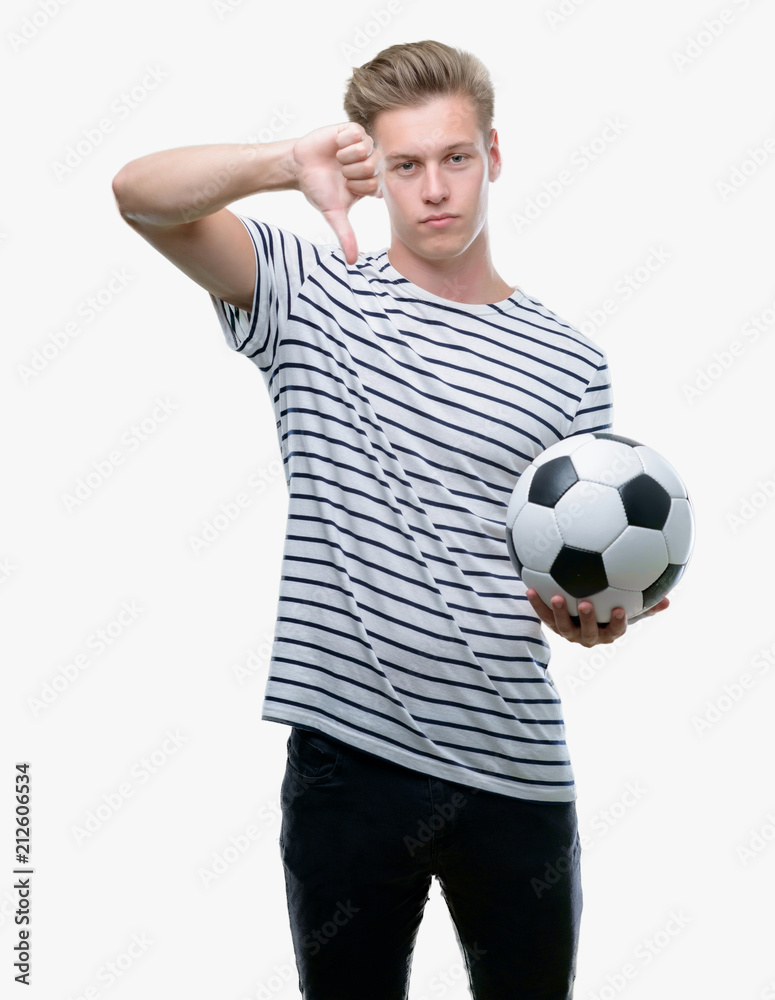Young handsome blond man holding soccer ball with angry face, negative sign showing dislike with thumbs down, rejection concept Young handsome blond man holding soccer ball with angry face, negative sign showing dislike with thumbs down, rejection concept