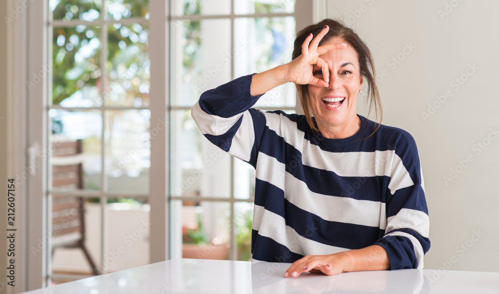 Middle aged woman with happy face smiling doing ok sign with hand on eye looking through fingers Middle aged woman with happy face smiling doing ok sign with hand on eye looking through fingers