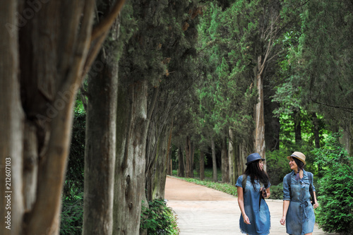 Two young beautiful brunette girls in jeans dresses and hats walk along the road of park with tall green trees.