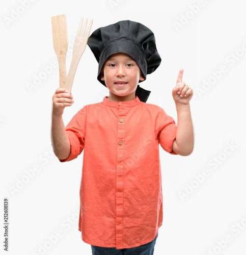 Dark haired little child wearing chef uniform surprised with an idea or question pointing finger with happy face, number one