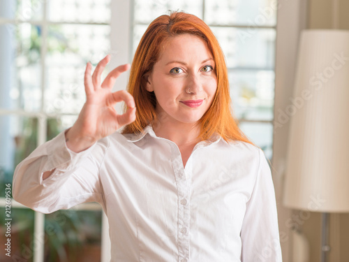 Redhead woman wearing white shirt at home doing ok sign with fingers, excellent symbol
