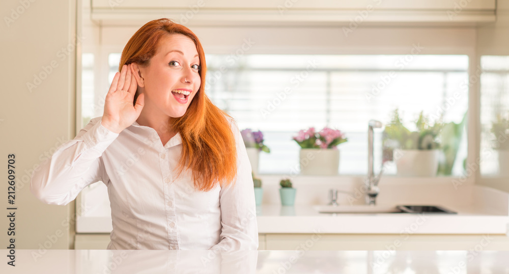 Redhead woman at kitchen smiling with hand over ear listening an hearing to rumor or gossip. Deafness concept.