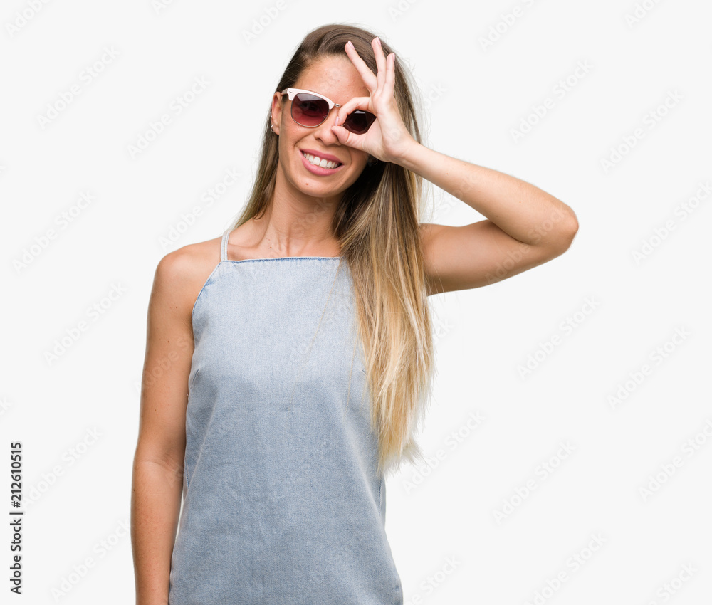 Beautiful young woman wearing sunglasses and denim dress with happy face smiling doing ok sign with hand on eye looking through fingers Beautiful young woman wearing sunglasses and denim dress with happy face smiling doing ok sign with hand on eye looking through fingers