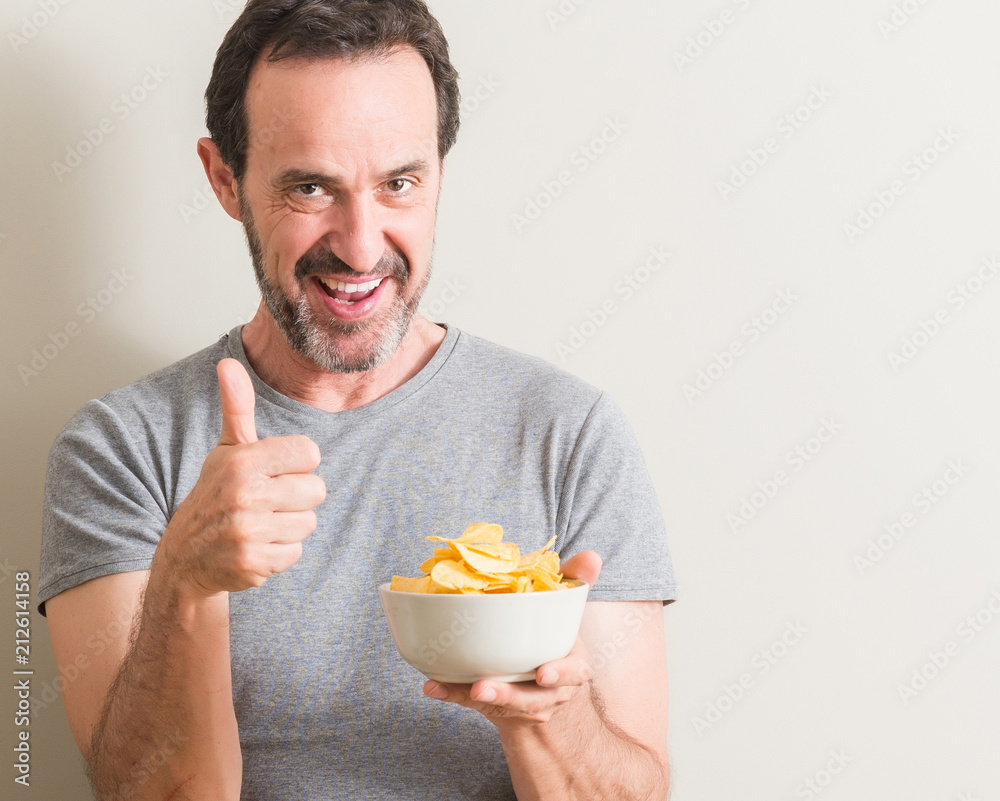 Senior man eating potato chips happy with big smile doing ok sign, thumb up with fingers, excellent sign