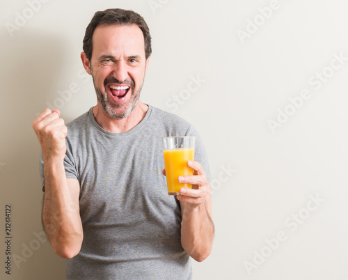 Senior man drinking orange juice in a glass screaming proud and celebrating victory and success very excited, cheering emotion