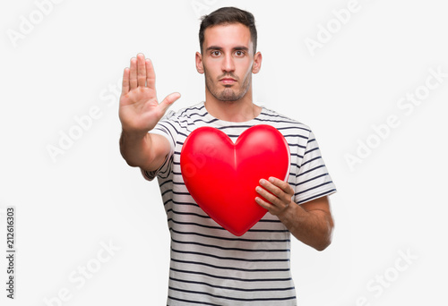 Handsome young man in love holding red heart with open hand doing stop sign with serious and confident expression, defense gesture © Krakenimages.com