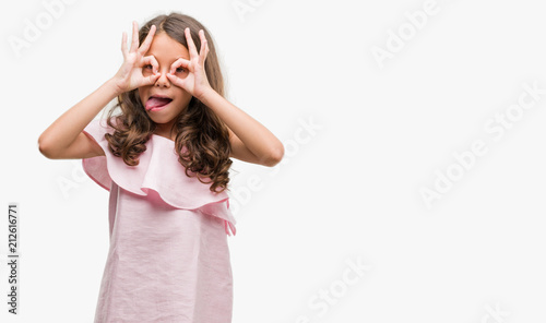 Brunette hispanic girl wearing pink dress doing ok gesture like binoculars sticking tongue out, eyes looking through fingers. Crazy expression.