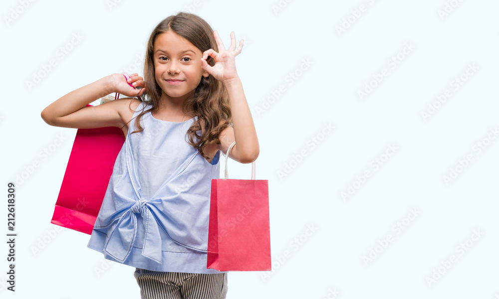 Brunette hispanic girl holding shopping bags doing ok sign with fingers, excellent symbol