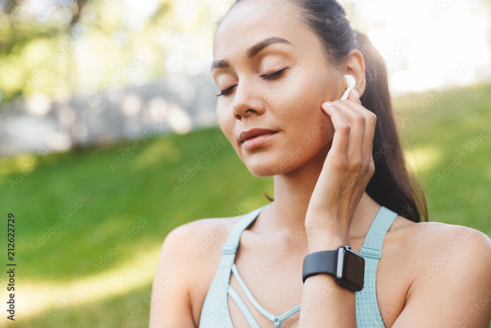 Image closeup of pretty satisfied woman 20s in sportswear using bluetooth earphone and listening to music with closed eyes, during walk in green park Image closeup of pretty satisfied woman 20s in sportswear using bluetooth earphone and listening to music with closed eyes, during walk in green park