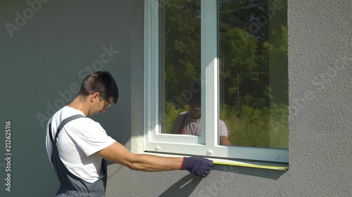 Male worker in safety glasses and protective gloves measuring sill and PVC window frame size. Builder checking components fitting of construction outside building. Technology, exterior design concept