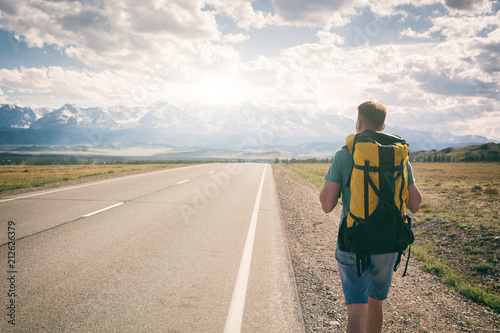 Man walks along an asphalt road with a backpack on his back overlooking the mountains