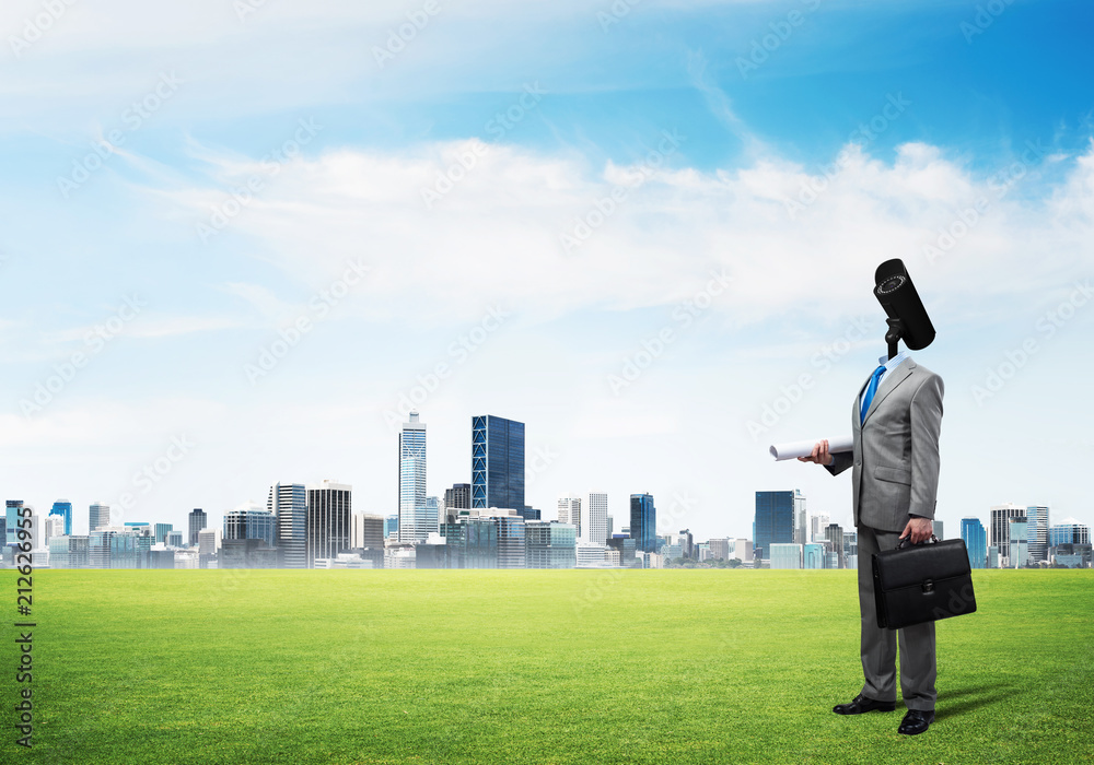 Camera headed man standing on green grass against modern cityscape Camera headed man standing on green grass against modern cityscape