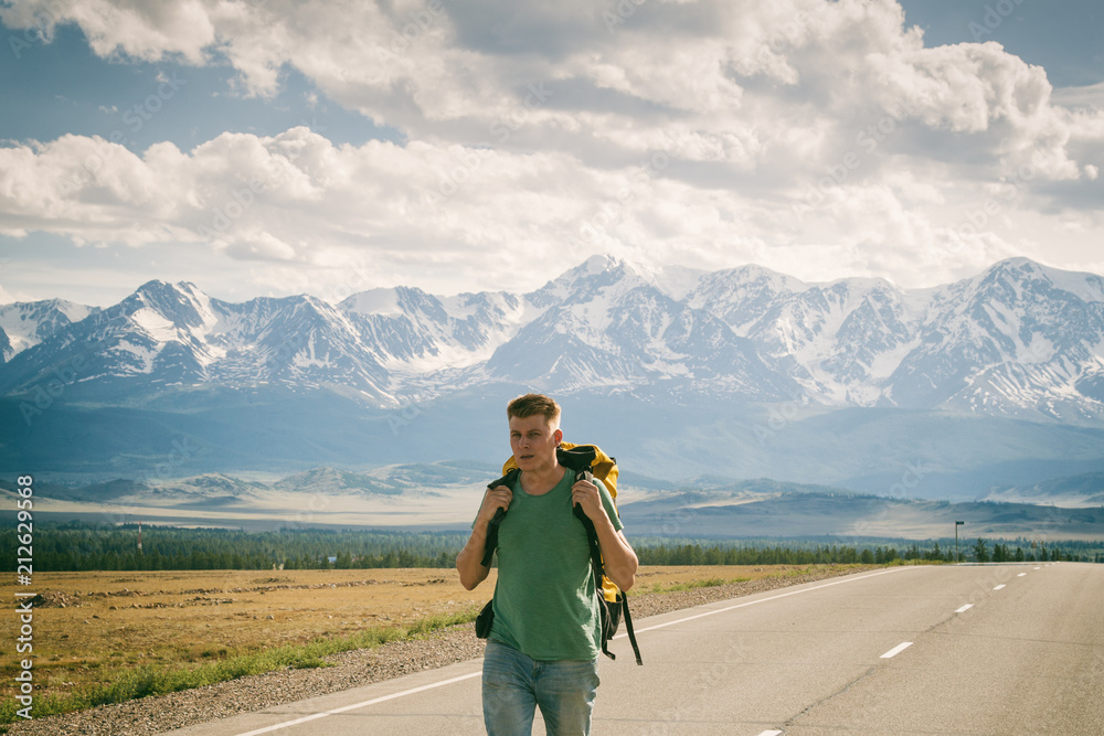 Man walks along an asphalt road with a backpack on his back. Mountain background